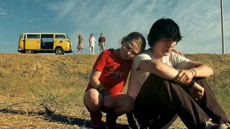 An screenshot of the movie Little Miss Sunshine, as a boy is upset and sits in a field while his sister comforts him, with their family watching in the distance.